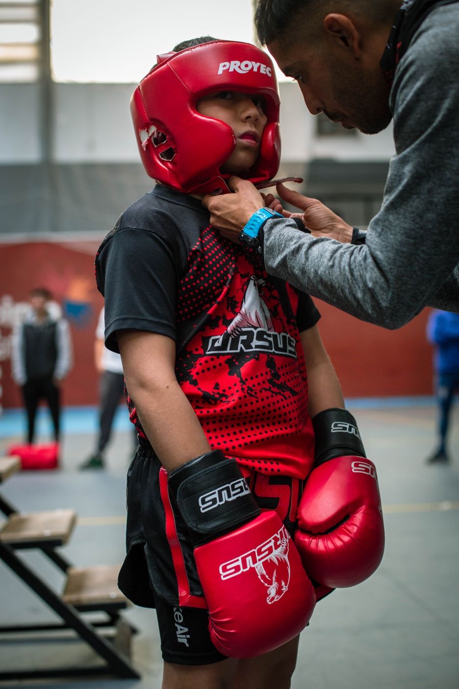 A boxing coach fastening the headgear of a young boxer in red gloves