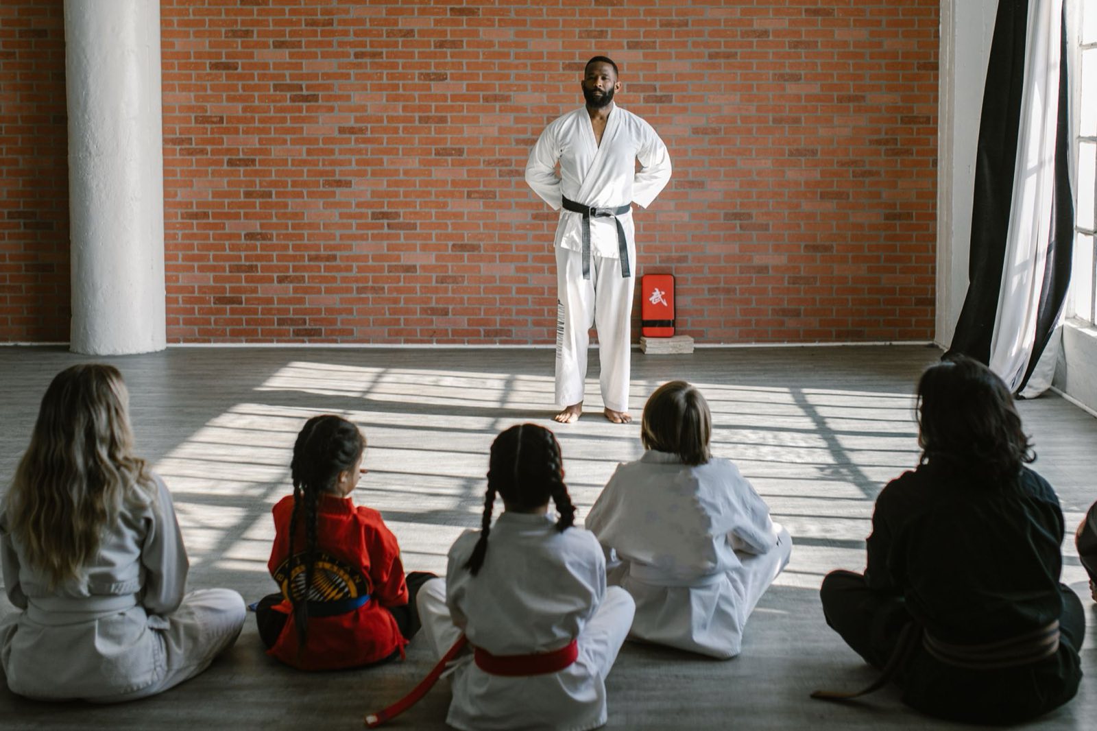 A karate instructor in a black belt leading a class of children and adults seated on the dojo floor