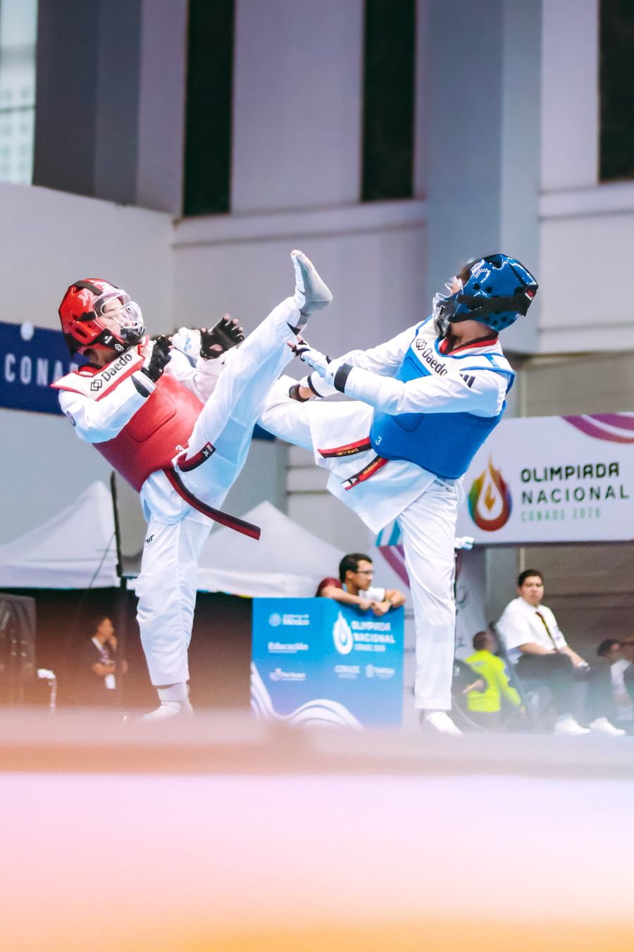 Two Taekwondo athletes in chest protectors and headgear trading high kicks in a match